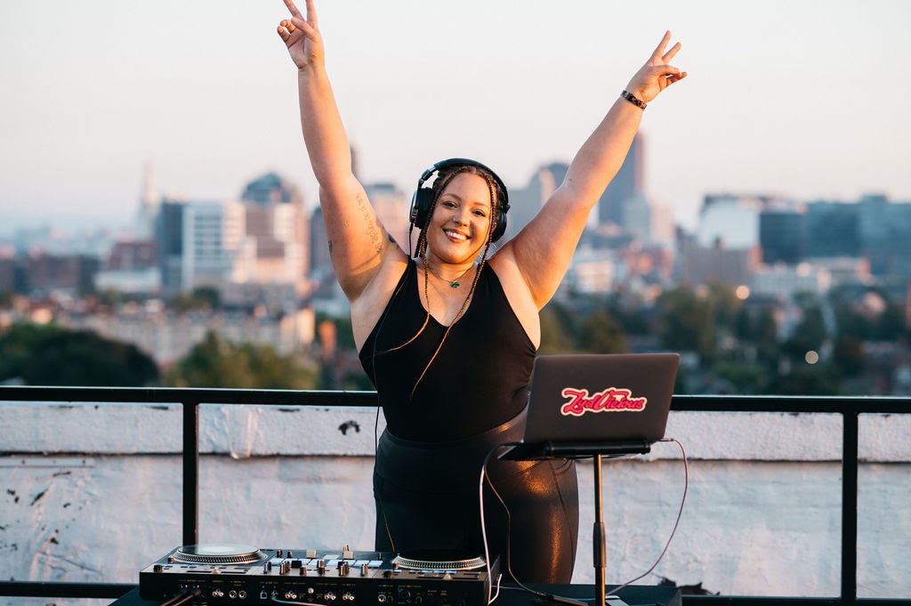 Lydia standing behind the DJ equipment with her arms up in a victorious pose. The Buffalo, NY skyline is behind her.